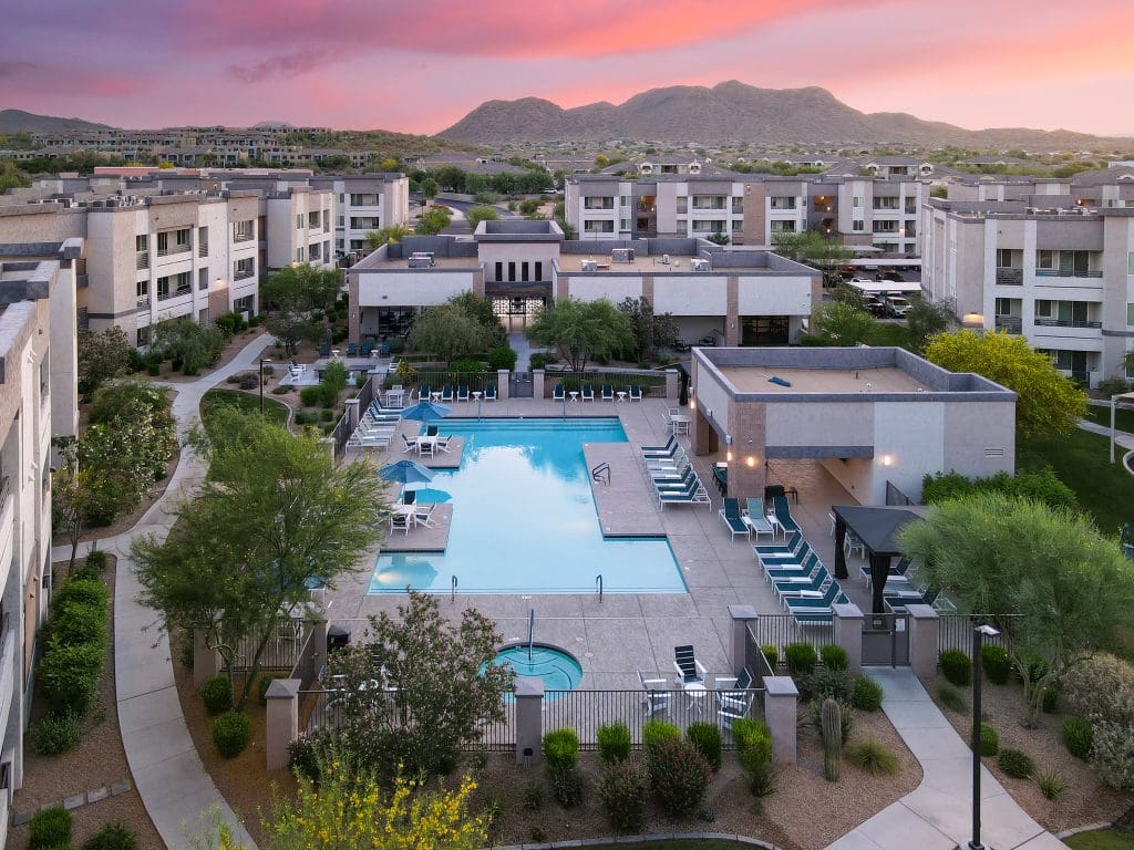 A large outdoor swimming pool with lounge chairs and landscaping is surrounded by modern apartment buildings, with mountains visible in the background at sunset.