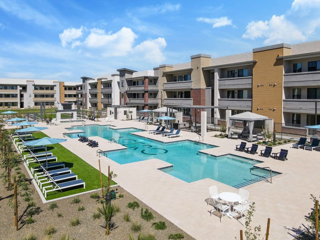 Modern apartment complex with three-story buildings, a large outdoor swimming pool, lounge chairs, umbrellas, and landscaped areas under a partly cloudy sky.