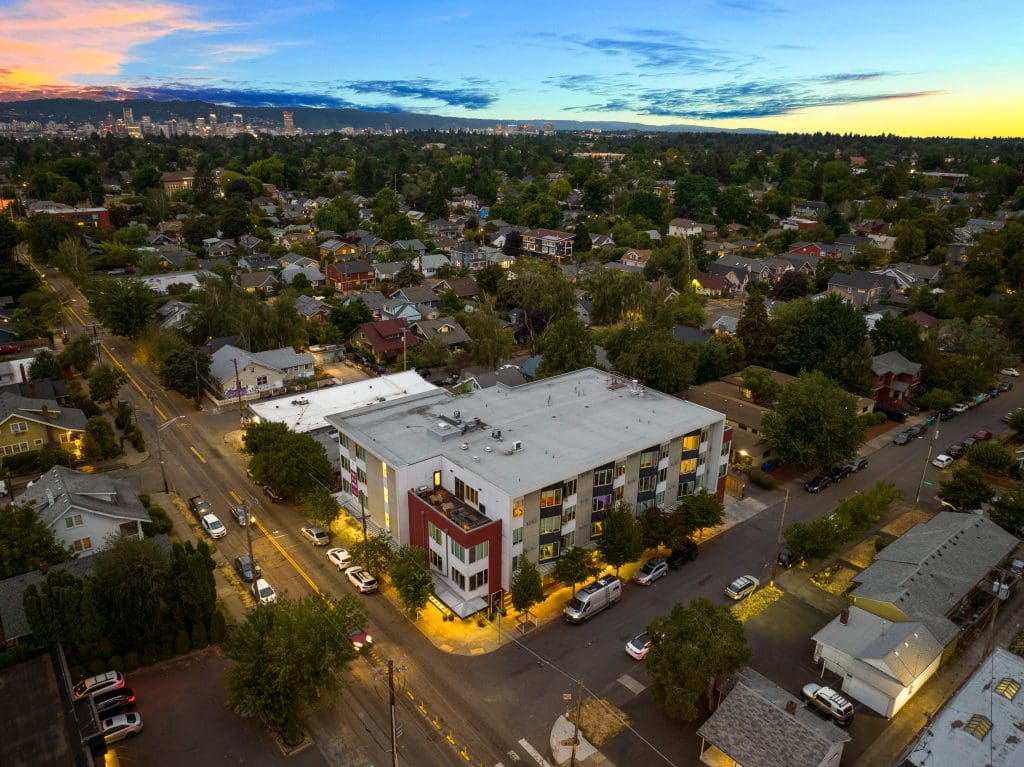 Aerial view of a mid-rise apartment building at dusk, surrounded by residential houses and tree-lined streets, with a city skyline visible in the distance.