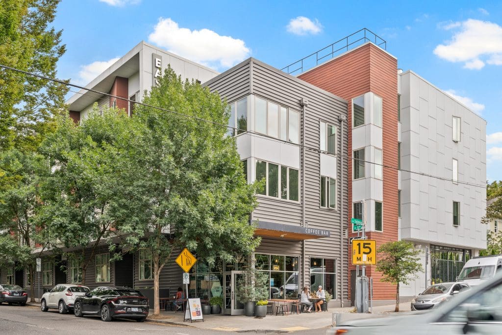 Modern mixed-use building with large trees, a coffee bar on the ground floor, parked cars, and a 15 mph speed limit sign on a sunny day.