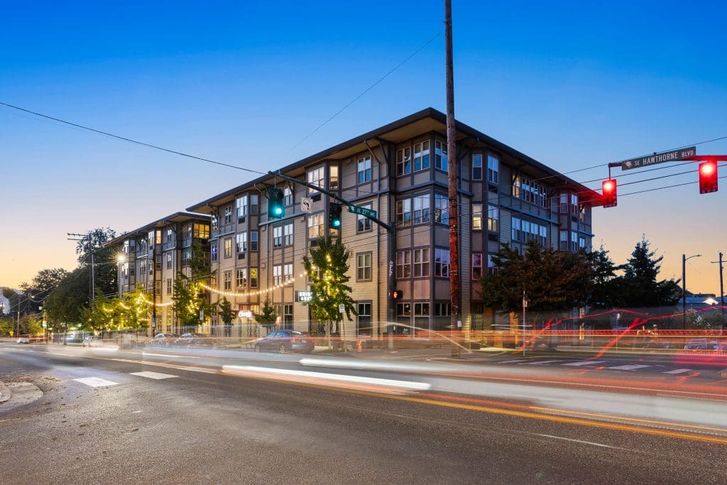 Four-story apartment building at an intersection with moving traffic and illuminated streetlights during dusk.