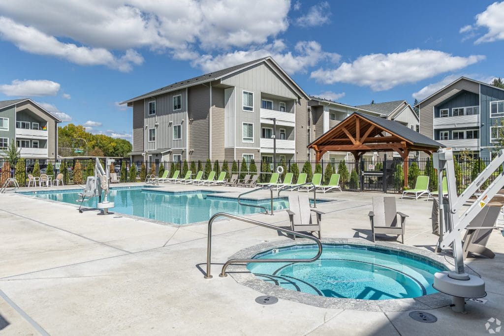 Outdoor swimming pool and hot tub area with lounge chairs, a wooden gazebo, and multi-story apartment buildings in the background under a partly cloudy sky.