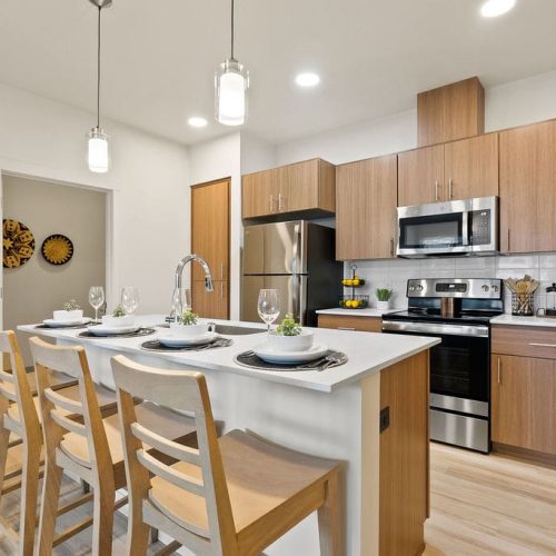 Modern kitchen with wooden cabinets, stainless steel appliances, island with sink, and three wooden barstools set with plates and glasses. Pendant lights hang above the island.