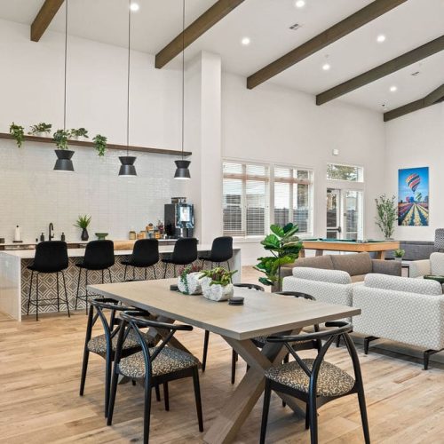 Modern communal lounge with a kitchen, dining table, bar seating, pool table, and lounge chairs. Large windows, plants, and wall art create a bright, inviting atmosphere.