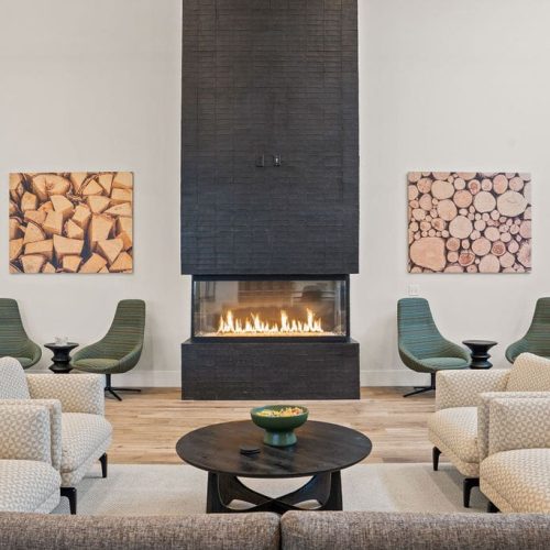 Modern living room with a central black fireplace, neutral armchairs, green accent chairs, wood-themed wall art, and a round black coffee table.