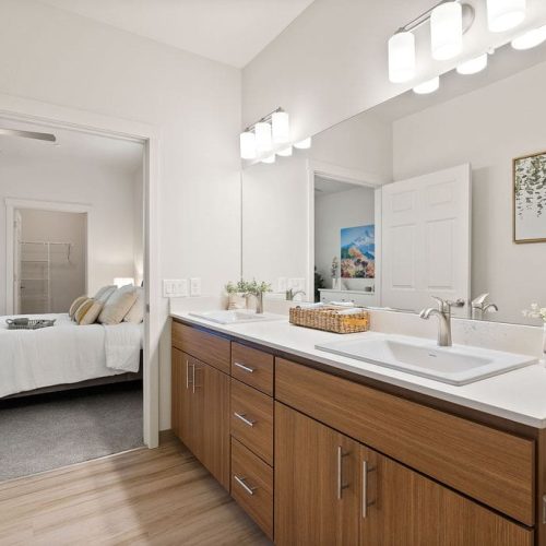 Modern bathroom with wooden cabinets, white countertop, rectangular sink, wall-mounted lights, and decorative plants; open door reveals a bedroom in the background.