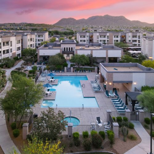 A large outdoor swimming pool with lounge chairs and landscaping is surrounded by modern apartment buildings, with mountains visible in the background at sunset.
