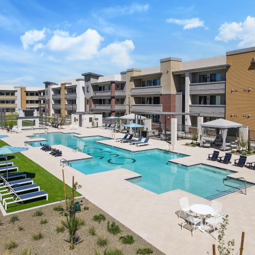 Modern apartment complex with three-story buildings, a large outdoor swimming pool, lounge chairs, umbrellas, and landscaped areas under a partly cloudy sky.