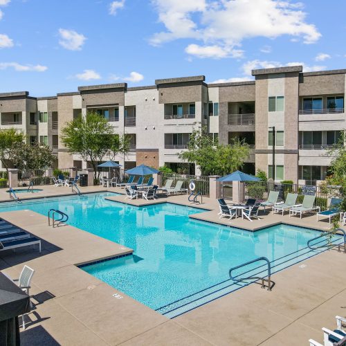 Outdoor swimming pool with lounge chairs and umbrellas surrounded by a modern three-story apartment complex under a clear sky.