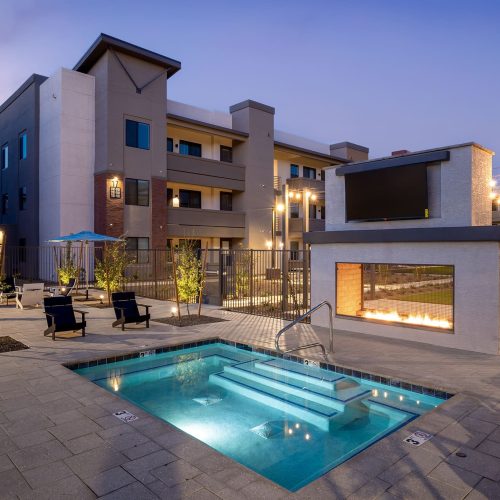 Outdoor pool and lounge area with fire feature, flat-screen TV, seating, string lights, and modern apartment buildings in the background at dusk.