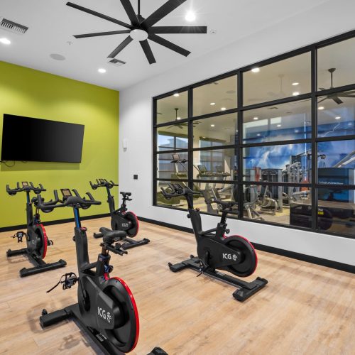 A modern indoor cycling room with four stationary bikes, wood flooring, a wall-mounted TV, and large glass windows overlooking a gym area.