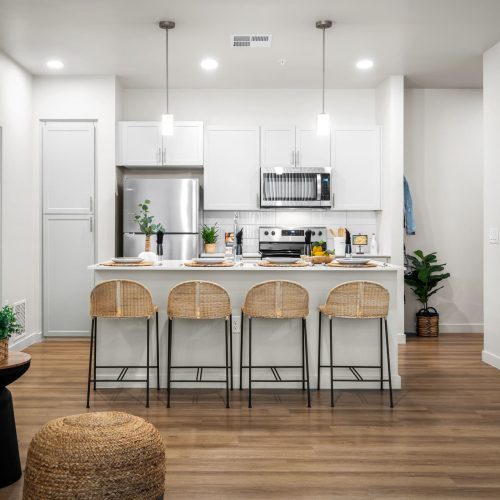 Modern kitchen with white cabinets, stainless steel appliances, a center island with four wicker barstools, pendant lights, and floral artwork on the wall.