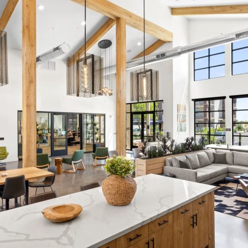 Modern open-concept lounge with high ceilings, large windows, exposed beams, a gray sectional sofa, assorted seating, and a kitchen island with plants and wooden decor.