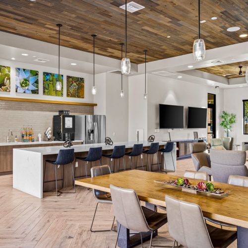 Modern communal kitchen and lounge area with bar seating, dining table, contemporary decor, large windows, and wall-mounted TVs.