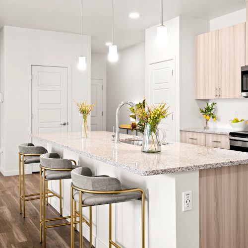 Modern kitchen with a large island, three barstools, stainless steel appliances, white cabinets, and pendant lighting. Yellow flowers in vases decorate the countertop.