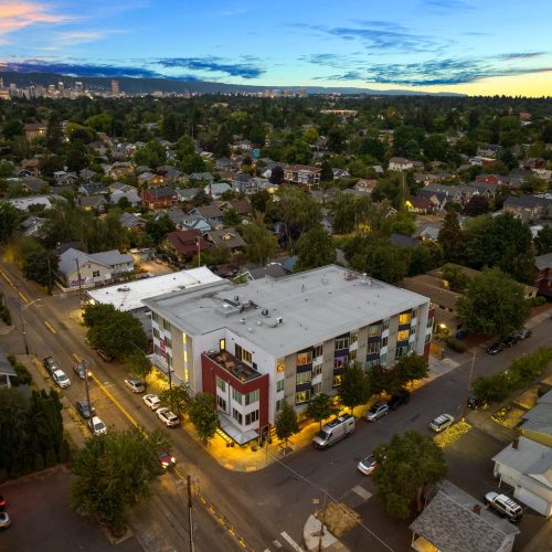 Aerial view of a mid-rise apartment building at dusk, surrounded by residential houses and tree-lined streets, with a city skyline visible in the distance.