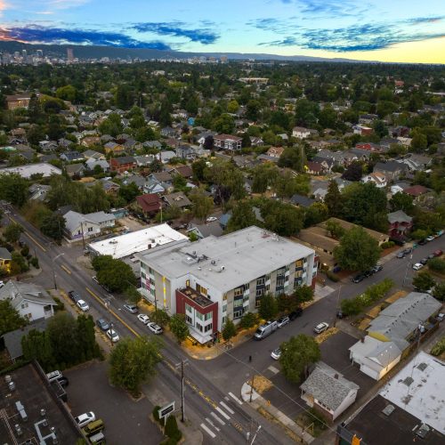 Aerial view of a residential neighborhood with a large apartment building at a street intersection, surrounded by houses, trees, and parked cars, under a partly cloudy sky at sunset.