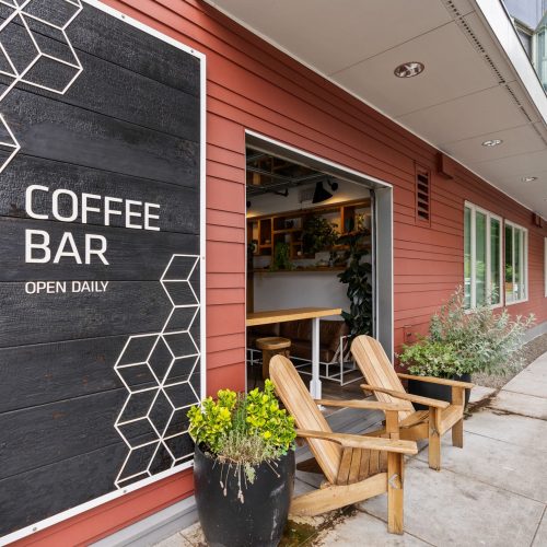 Exterior view of a coffee bar with a large sign, wooden chairs, potted plants, and an open window counter along a sidewalk.