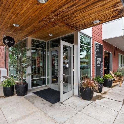 Glass door entrance to a modern coffee bar with potted plants and a wooden awning, located on a tree-lined sidewalk corner.