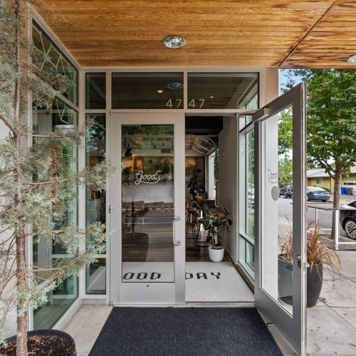 Glass entrance of a building with "Good Day" written on the floor mat, an open door, potted plants, and the number 4747 above the door. A street with cars and houses is visible outside.
