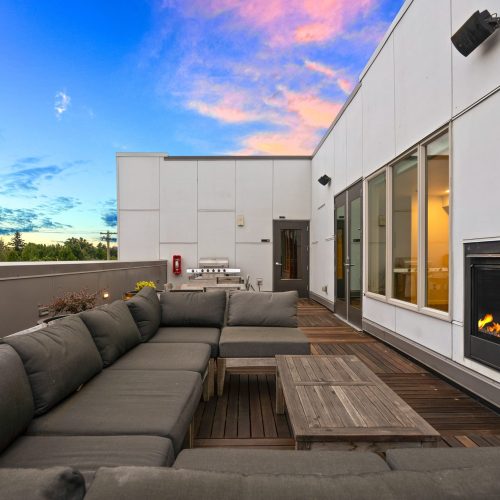 Modern rooftop patio with gray sectional sofas, wooden tables, a built-in fireplace, and a sunset sky in the background.