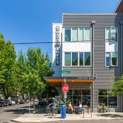 A modern apartment building with a "East Lofts" sign, a coffee bar at street level, and people sitting outside on a sunny day at a city intersection.