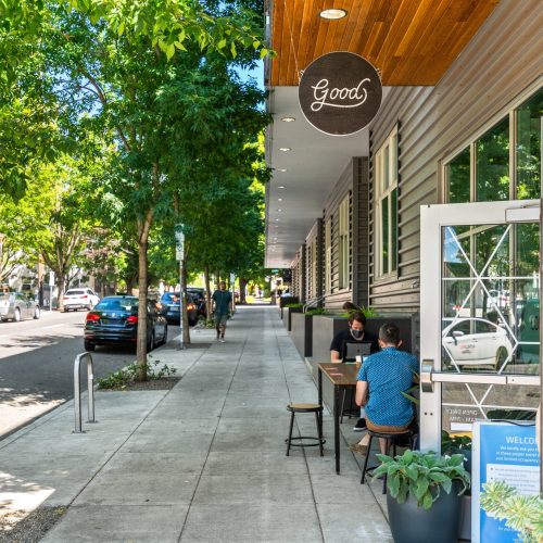 Two people sit at a table outside a café on a tree-lined city sidewalk with parked cars and greenery on a sunny day.
