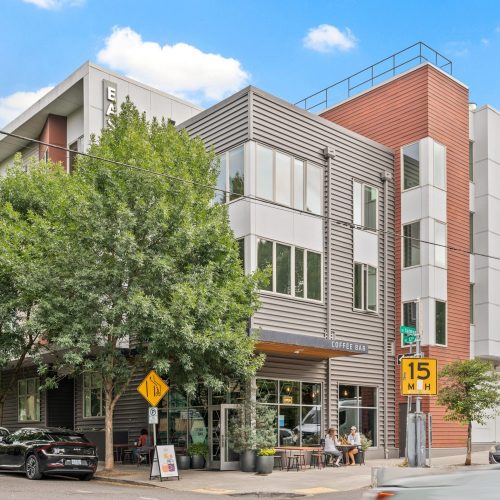 Modern mixed-use building with large trees, a coffee bar on the ground floor, parked cars, and a 15 mph speed limit sign on a sunny day.