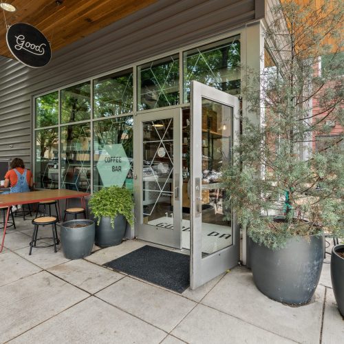 A modern coffee bar with large windows, outdoor seating, potted plants, and a glass door open to the sidewalk. One person is sitting at a table.