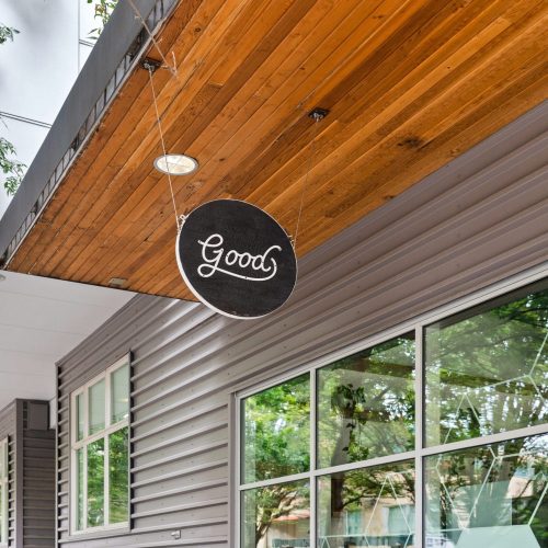A round sign with the word “Good” hangs from a wooden overhang outside a modern building with large windows and metal siding.