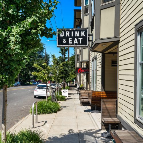A sidewalk scene shows a "DRINK & EAT" sign outside a restaurant with benches, trees, and parked cars along a sunny street.