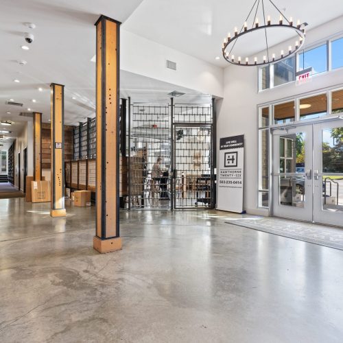A modern building lobby with polished concrete floors, wooden beams, a circular chandelier, large windows, and a gated area leading to an interior space.