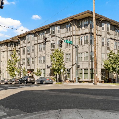 Four-story modern apartment building on a street corner, with parked cars along the curb and trees lining the sidewalk under a blue sky.