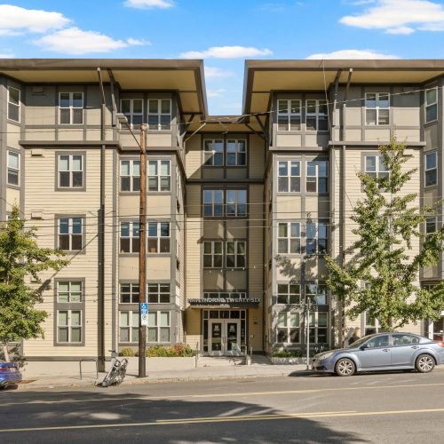 Four-story beige apartment building with multiple windows, a central glass entrance, and cars parked along the street in front.