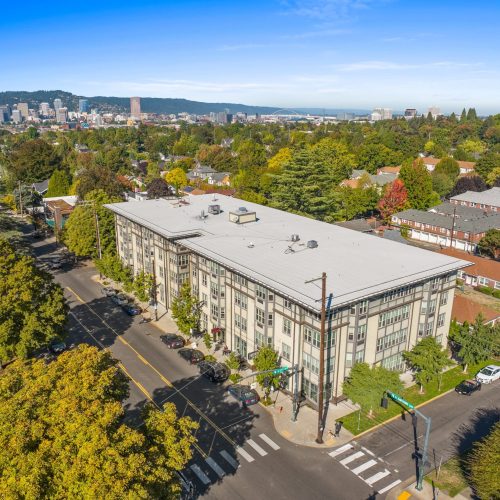 Aerial view of a large rectangular building with a flat roof, surrounded by trees and residential streets, with a city skyline visible in the background.