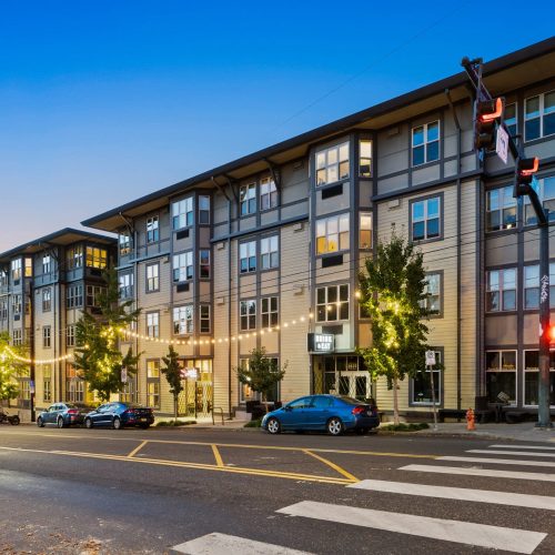 A modern four-story apartment building with large windows, street-level businesses, parked cars, trees, and a crosswalk at a city intersection during dusk.