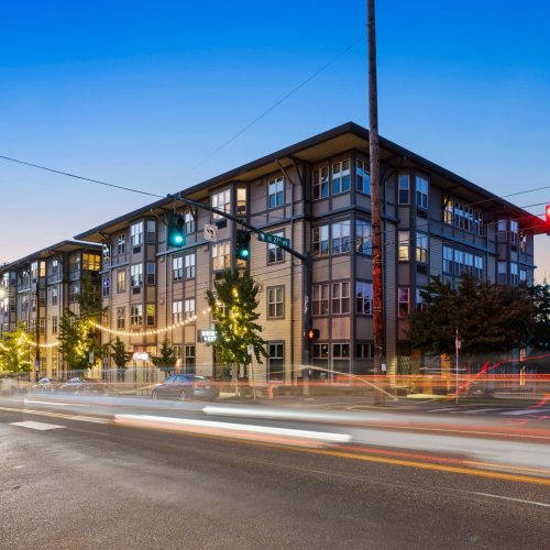 Four-story apartment building at an intersection with moving traffic and illuminated streetlights during dusk.