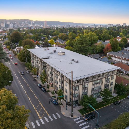 Aerial view of a mid-rise apartment building at a street intersection in a leafy residential neighborhood with a city skyline in the background.