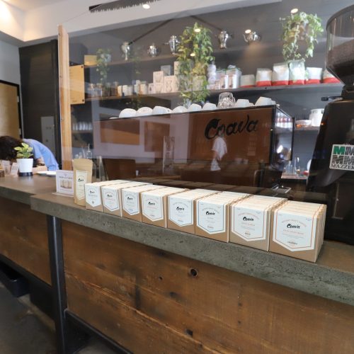 A countertop at a coffee shop displays several boxes of coffee in a row, with plants and coffee equipment visible in the background.