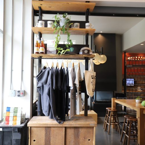 A retail display inside a café shows hanging t-shirts, mugs, bottles, and a potted plant on wooden shelves, with tables and stools in the background.