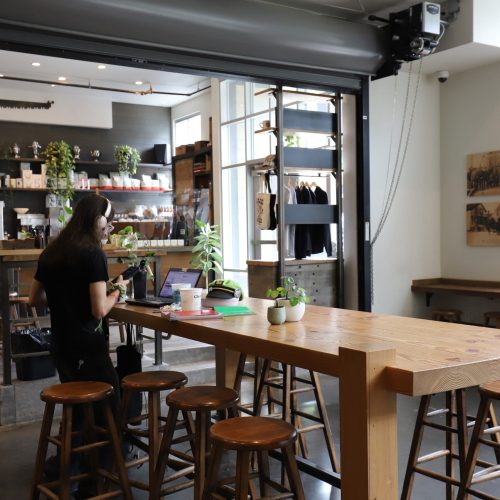 A person sits at a wooden table using a laptop in a modern café with plants, shelves, stools, and natural light from the windows.