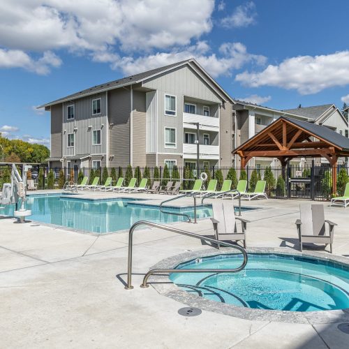 Outdoor swimming pool and hot tub area with lounge chairs, a wooden gazebo, and multi-story apartment buildings in the background under a partly cloudy sky.