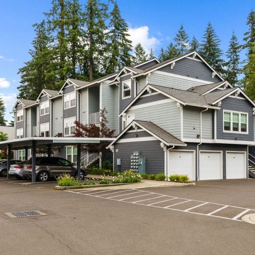 A modern multi-unit apartment building with gray siding, white trim, attached garages, and a covered parking area, surrounded by trees and a paved driveway.