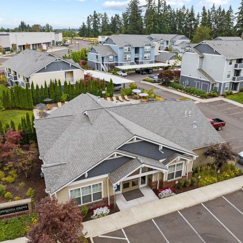 Aerial view of a suburban apartment complex with multiple gray buildings, landscaped greenery, parking spaces, and sidewalks.