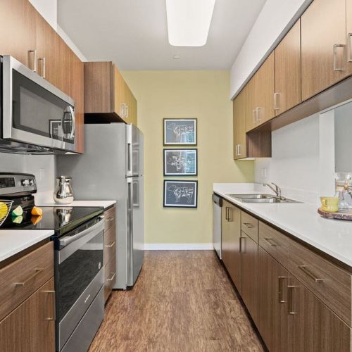 Modern kitchen with wood cabinets, stainless steel appliances, white countertops, and a sink. Artwork and a TV are visible on the yellow back wall and right counter.