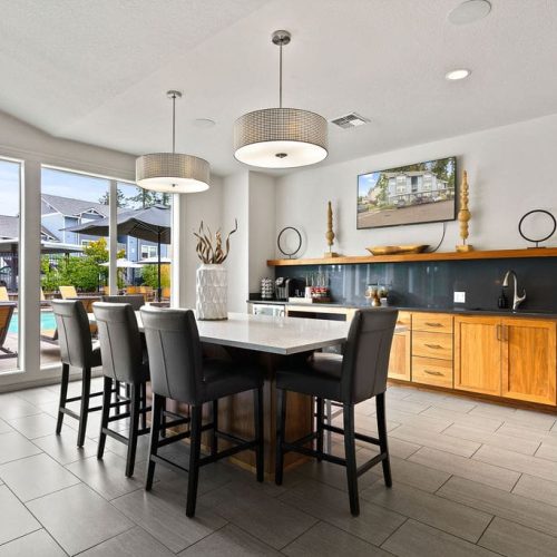 Modern kitchen with a central island, black chairs, wooden cabinets, stainless steel appliances, and large windows overlooking a pool and outdoor seating area.