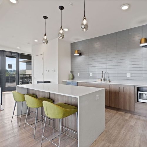 Modern kitchen with a large island, three green barstools, pendant lighting, wood flooring, built-in microwave, two decorative vases, and a seating area by large windows.