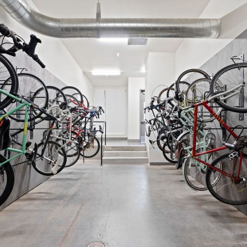 A clean indoor bike storage room with bicycles mounted on wall racks along both sides, concrete floors, and exposed ductwork on the ceiling.