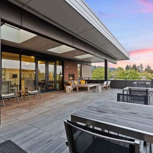 Spacious rooftop patio with wooden decking, outdoor seating, and tables overlooking greenery, under a colorful sunset sky.